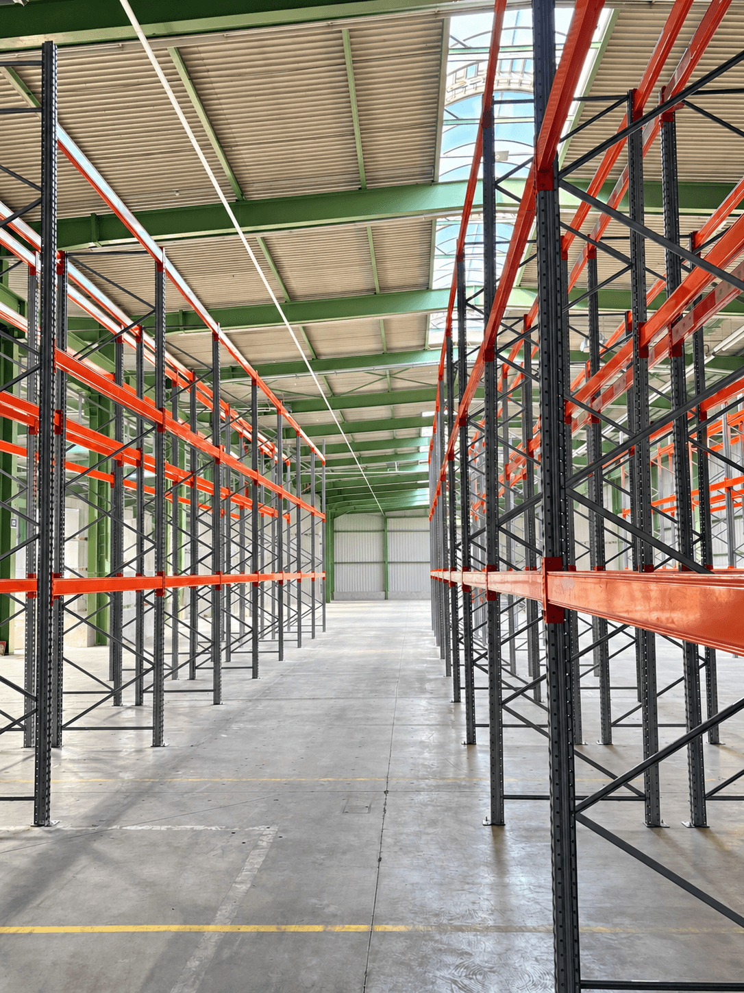Blick durch eine leere Lagerhalle mit hohen, orangefarbenen Schwerlastregalen und hellem Tageslicht von oben.