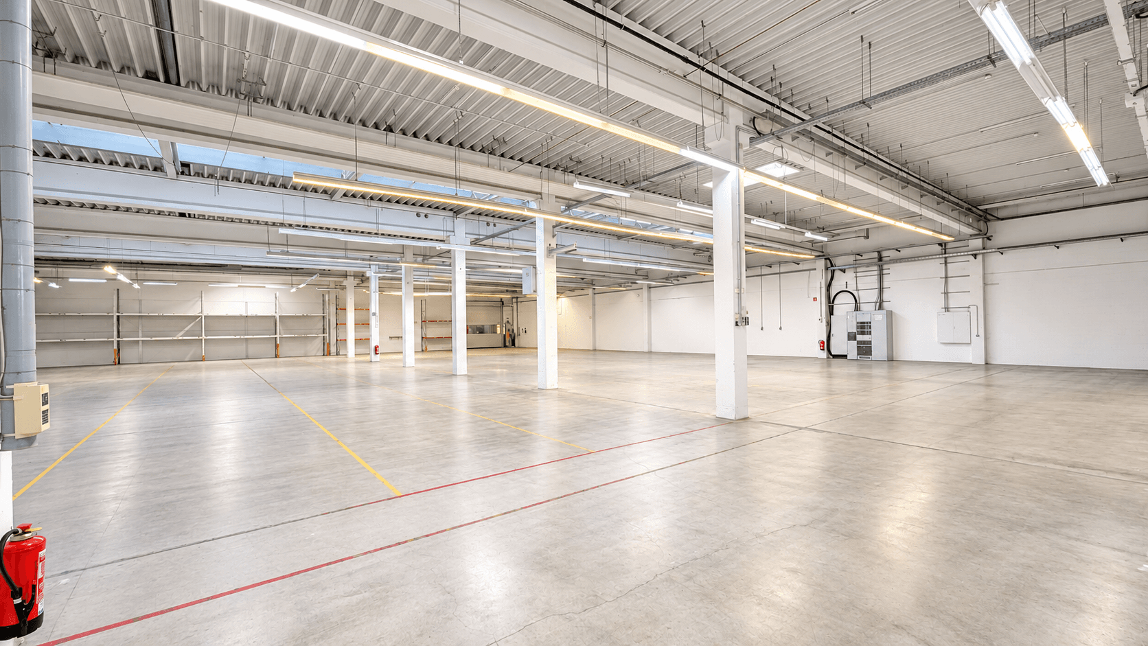 Helle, leere Lagerhalle mit weißen Säulen, Betonboden und moderner Deckenbeleuchtung in einem Industriegebäude.